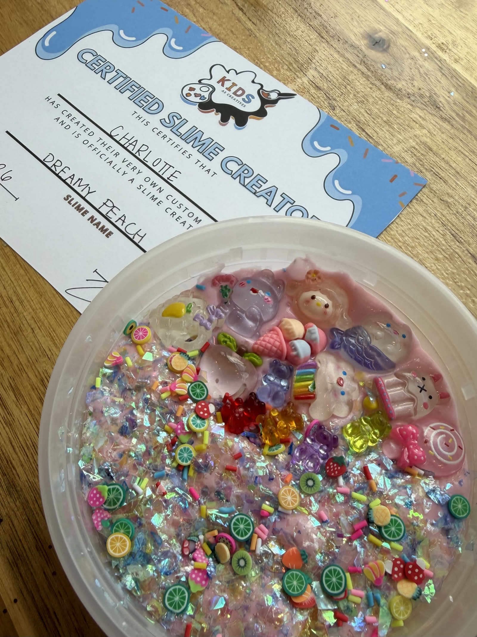 A custom slime filled with charms and glitter beside a Certified Slime Creator certificate