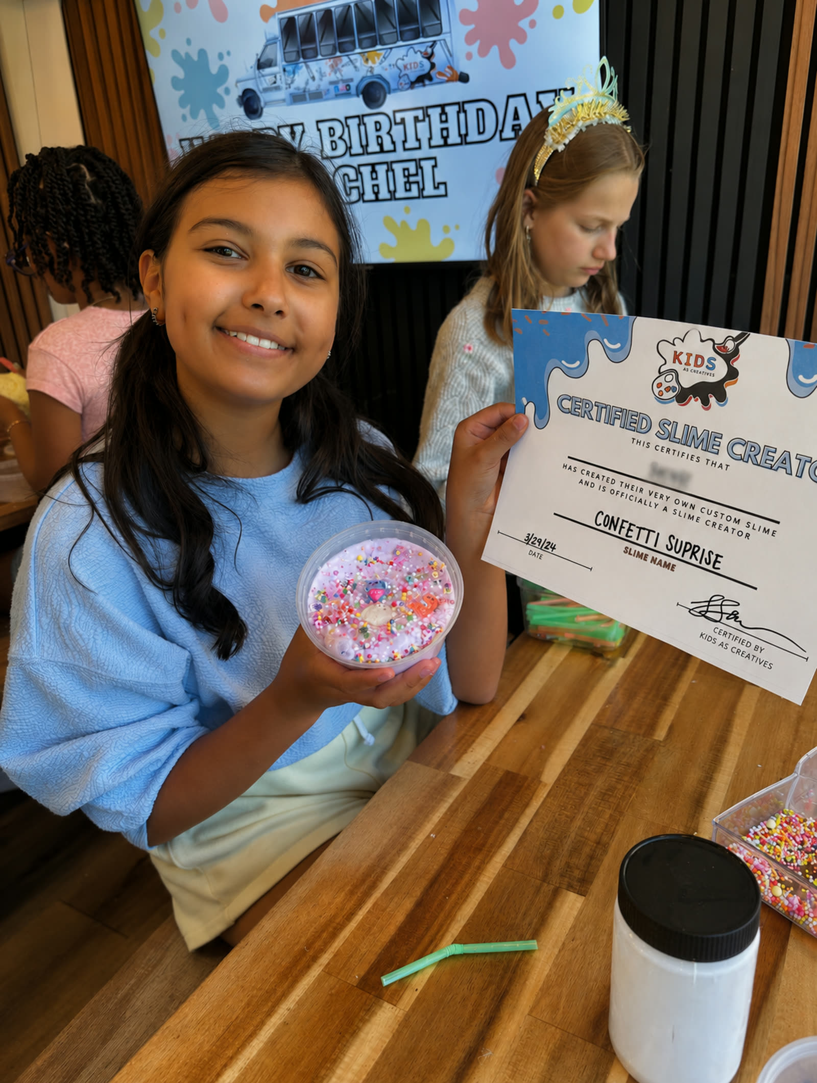 A smiling girl holding her custom confetti slime and a Certified Slime Creator certificate