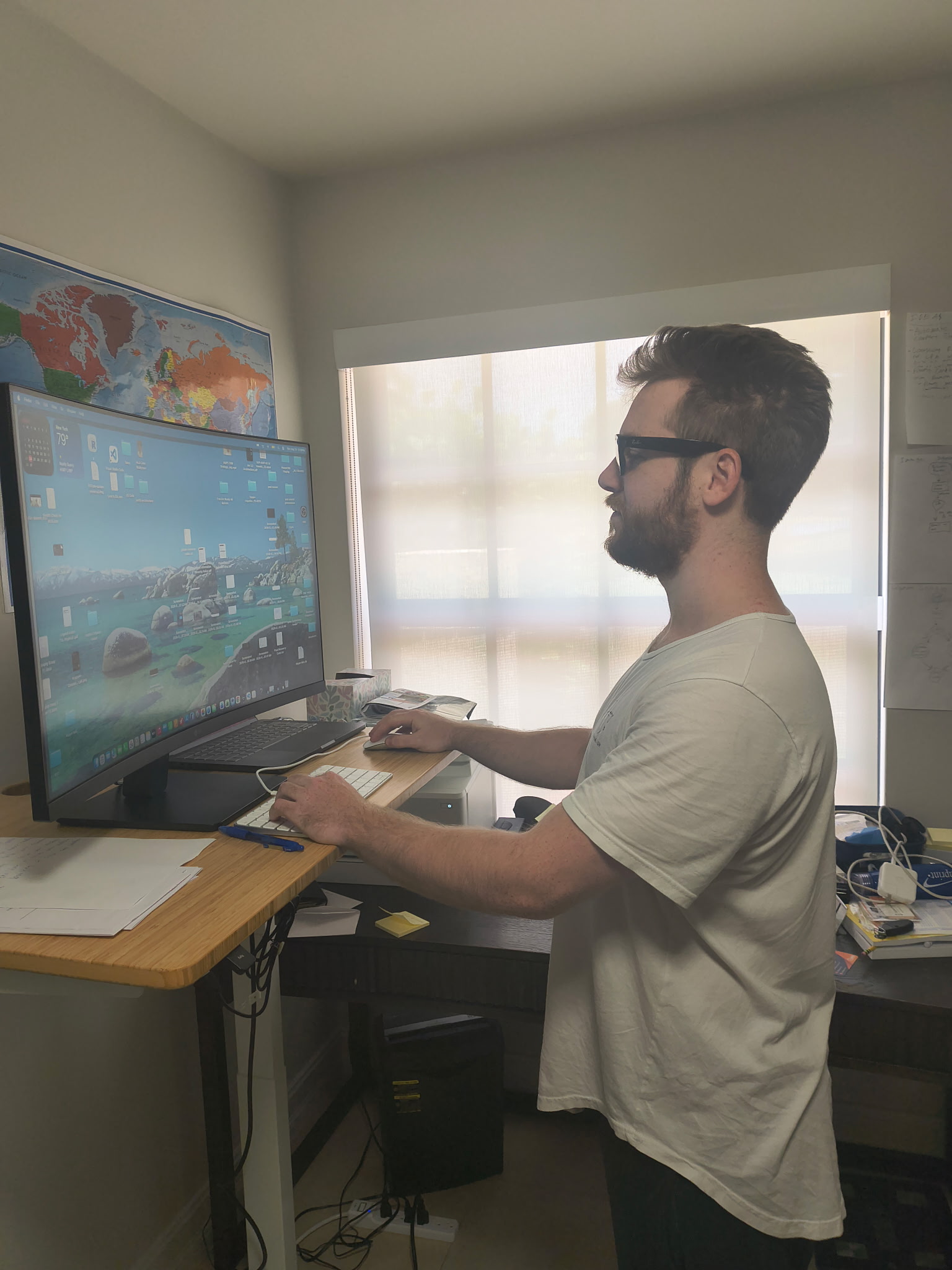 Joseph working at his new standing desk, standing at an elevated desk with a large monitor showing a nature wallpaper, a world map on the wall behind him