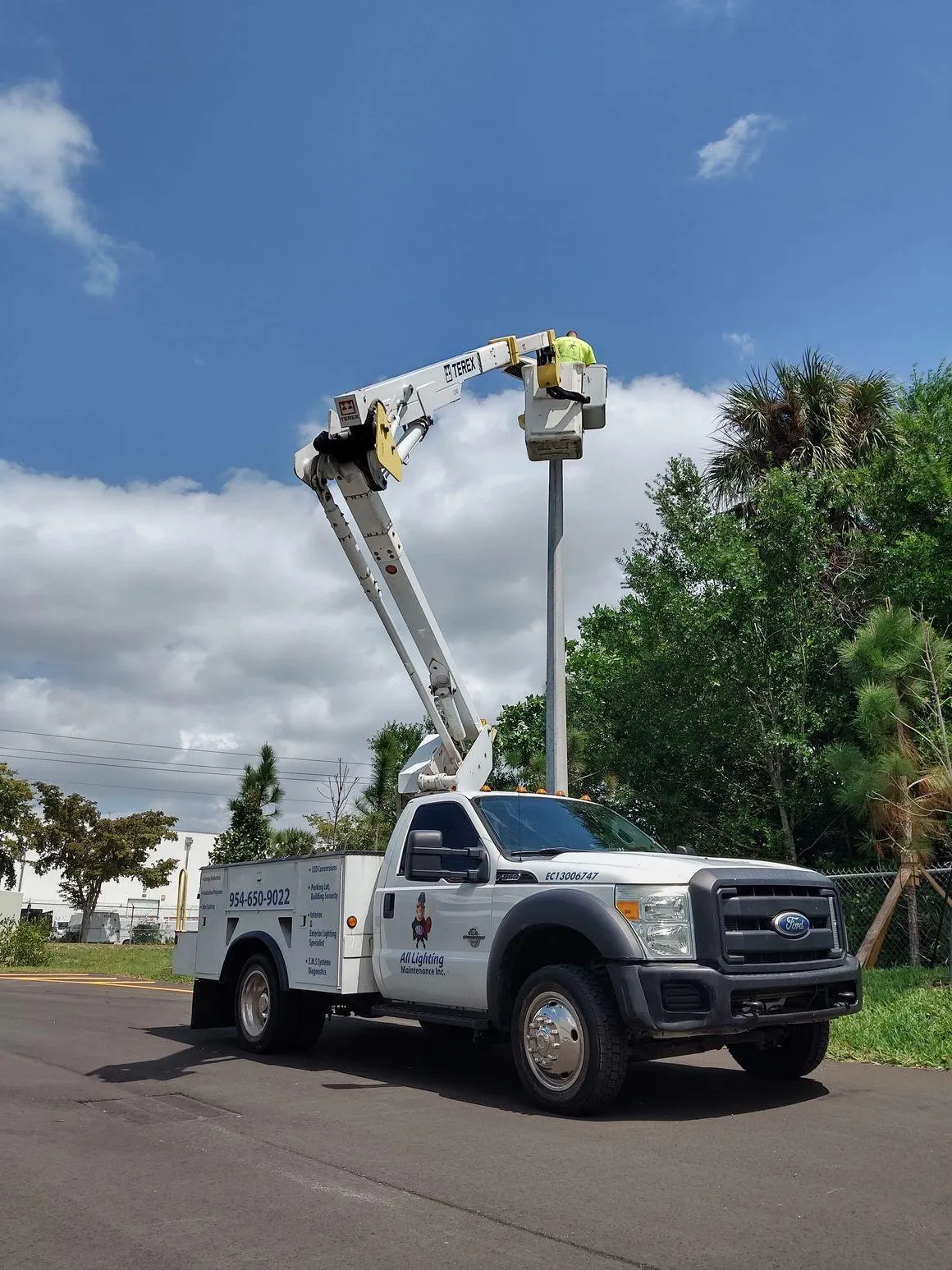 All Lighting Maintenance technician using a white Ford bucket truck to service a high-reach parking lot light pole.