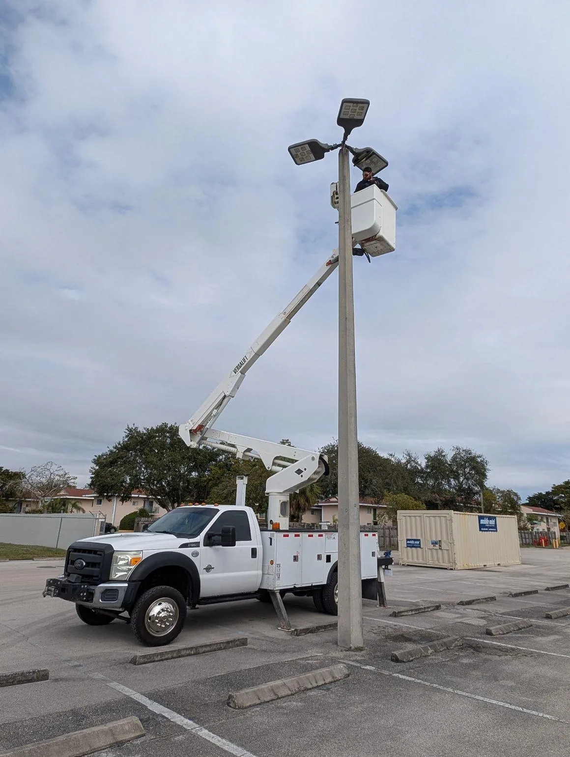 All Lighting Maintenance technician using a Versalift bucket truck to service a high-reach multi-head parking lot light pole.