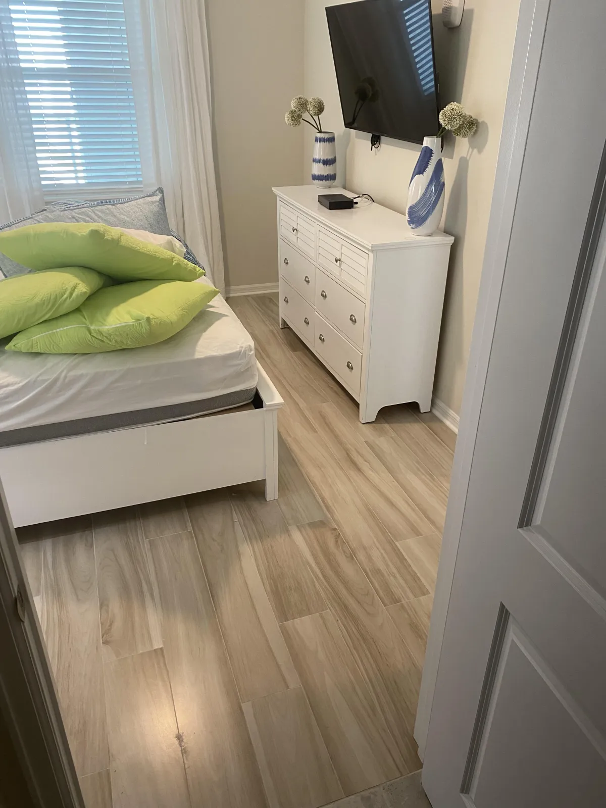 Modern bedroom featuring light oak wood-look plank flooring. A white dresser with a TV mounted above it sits against a light grey wall.