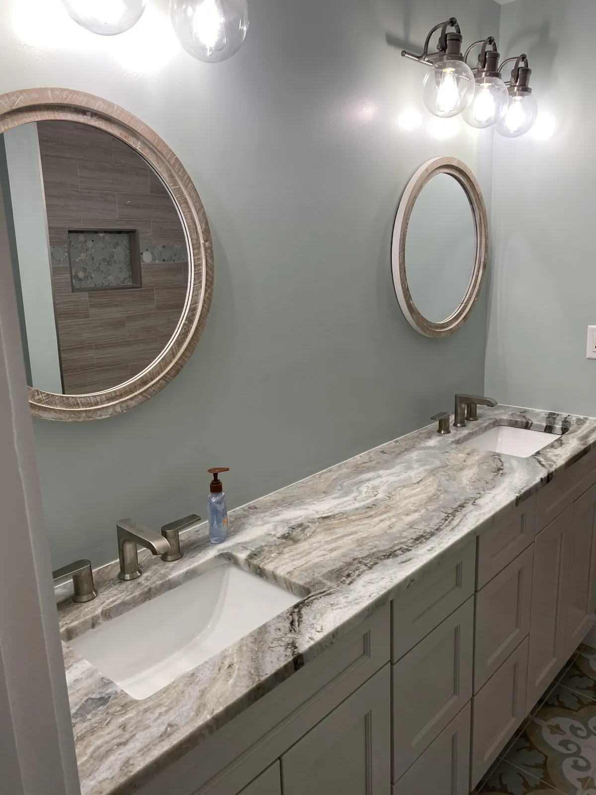 Master bathroom featuring a double vanity with grey and white natural stone countertops, white shaker cabinets, and round wood-framed mirrors.